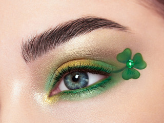 Conceptual photo of St. Patrick's Day. The eye of the young beautiful woman with bright green shadows and expressive eyebrows. Shamrock patterns. Holiday makeup - obrazy, fototapety, plakaty