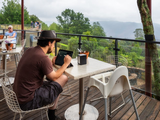 Young man drawing using his laptop, while relaxing at a cafe terrace after rain at Mount Tomah Botanic Garden in Blue Mountains National Park, NSW, Australia.