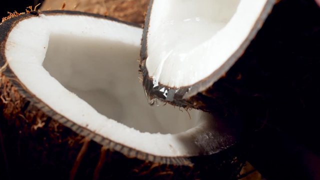 Slow motion closeup video of water slowly pouring from one coconut half into another part. Perfect abstract backdrop for organic food and healthy nutrition.