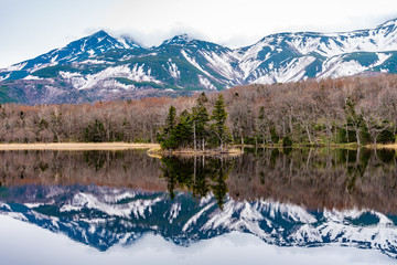 Small islet in the lake, beautiful lake surface reflecting blue sky like a mirror, rolling mountain range and woodland in the background on springtime sunny day. High latitude country natural scenery