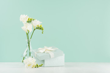 Festive background with opened square gift box and fresh white freesia flowers in elegant transparent vase on green mint menthe wall, white wood table. - obrazy, fototapety, plakaty