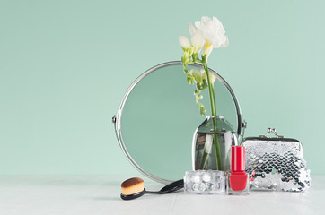 Dressing table in mint menthe with accessories for make up in silver with white flowers, round mirror, cosmetics bag, red nail polish on white wood.