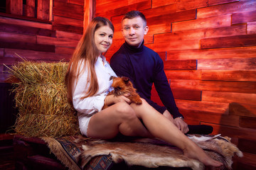 Obraz na płótnie Canvas Beautiful girl in white shirt and guy in black sweater with small dog on the couch with hay in the barn or in the hayloft with wooden walls. Couple in rustic interior during photo shoot