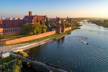 Fototapeta premium Medieval Malbork (Marienburg) Castle in Poland, main fortress of the Teutonic Knights at the Nogat river, bridges and a water scooter. Aerial view in sunset light.