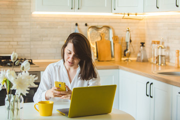 young pretty caucasian woman talking on the phone sitting in front of laptop in the kitchen