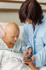 Woman without hair after chemotherapy patient lying at hospital bed feeling sad and depressed worried. A friend came to please her, showing photos on her mobile phone.