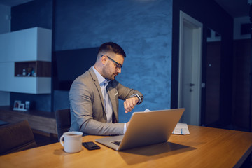 Side view of handsome unshaven serious businessman in suit and with eyeglasses sitting at dining table and looking at wristwatch. On table are laptop, coffee and smart phone. Morning time.