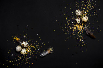 Moody easter background. Quail eggs, feathers and golden sugar sprinkles on black background. Happy...