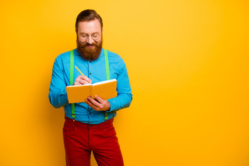 Photo of attractive guy holding writing notes in personal planner have creative idea for new project wear bright blue shirt green suspenders red pants isolated yellow color background