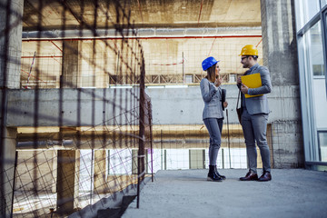 Full length of two colleagues with helmets on heads standing in building in construction process...