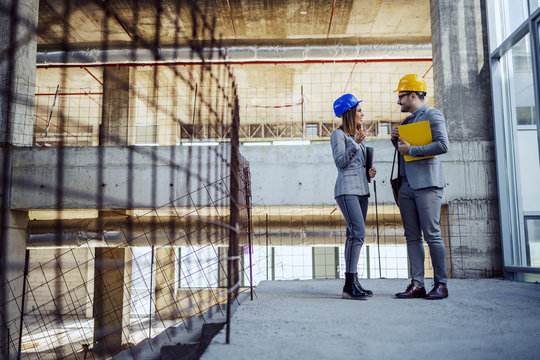 Full length of two colleagues with helmets on heads standing in building in construction process and talking about what they plan to change about project.
