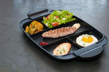 Breakfast, cutlet or steak, tomato salad, fried potatoes, croutons and fried egg on black plate. Restaurant lunch, content for the site