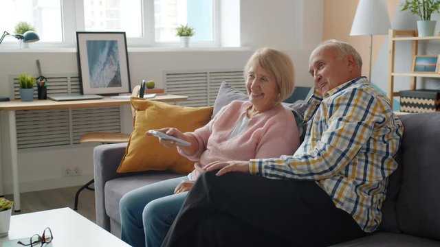 Senior man and woman watching TV and talking relaxing on couch in apartment having fun together. Modern technology and entertainment concept.