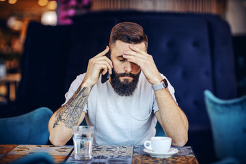 Young desperate caucasian bearded tattooed handsome hipster having a phone conversation and just receive bad news. Cafeteria interior.