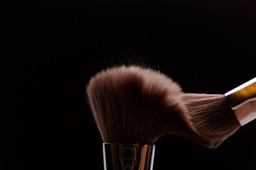 Make-up brush with beige powder explosion on black background. Creative splash. Professional powder. Cosmetics brush releasing a cloud of sparkling face powder. Copy space. Beauty and makeup concept