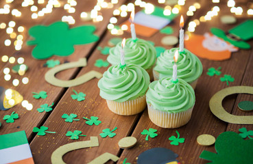 st patricks day, holidays and celebration concept - green cupcakes with candles, horseshoes and shamrock on wooden table over festive lights