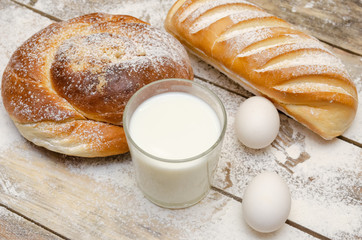 glass of milk, loaf, two eggs on a wooden background with flour