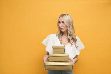 Happy blond european woman in white blouse holding a pack of gift boxes over yellow background. Copy space