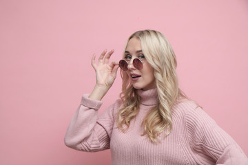 Happy coquettish woman face with blond hair in glasses and wool sweater on pastel pink background. Close up