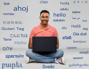 technology, communication and people concept - happy indian man with laptop computer sitting on floor over greeting words in different foreign languages on grey background