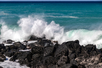 White wave breaking against black volcanic rocks  on the Kona coast of Hawaii's Big Island. Vivid blue-green pacific ocean in the distance.