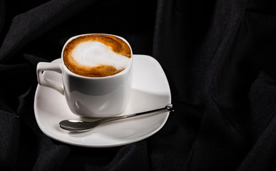 White ceramic cup of coffee with milk on small dish next to teaspoon, black fabric background