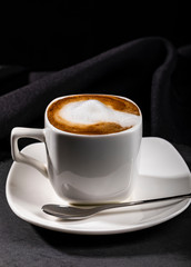 Close-up of ceramic cup of coffee with milk full to brim on white saucer next to teaspoon, low angle