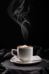 Steaming cup of coffee with milk froth on white saucer and board over dark fabric folds background