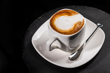 Close-up of cup of cappuccino on white squared saucer next to bizarre teaspoon, high angle, isolated