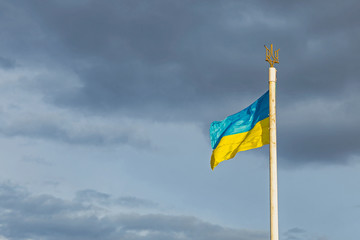 Flags and coat of arms of Ukraine on a flagpole against a cloudy sky. 