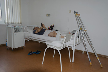 A man is lying in a Russian hospital after a traffic accident. He has a broken leg, his leg is plastered, and there are crutches nearby.