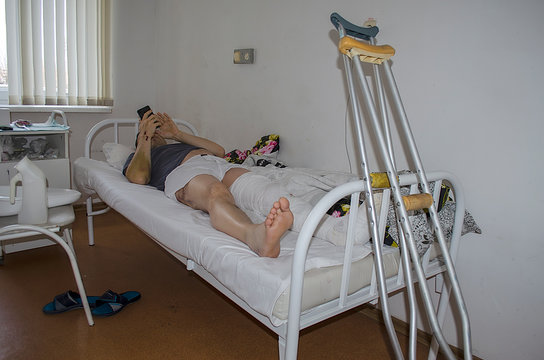 A Man Is Lying In A Russian Hospital After A Traffic Accident. He Has A Broken Leg, His Leg Is Plastered, And There Are Crutches Nearby. He Reads The News On His Phone.