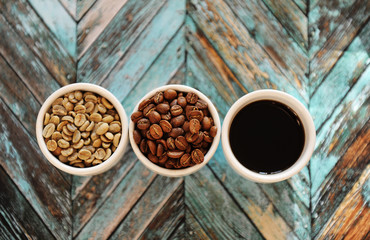 Green raw beans, roasted coffee beans and black coffee in three white cups on turquoise wooden background top view