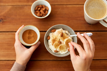 food and breakfast concept - hands of woman eating oatmeal porridge in bowl with sliced banana, almond nuts and cinnamon and cup of coffee on wooden table