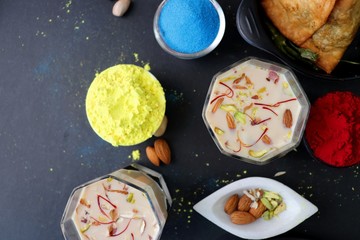 Assorted Indian Mahashivratri or Holi festival food like samosa, kachori, Kaju Katli, Puran Poli, dry fruits & Almond milk Thandai drink with Happy Holi wishes over black background with copy space.