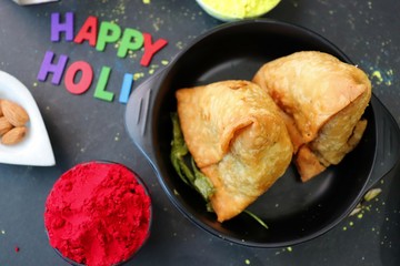 Assorted Indian Mahashivratri or Holi festival food like samosa, kachori, Kaju Katli, Puran Poli, dry fruits & Almond milk Thandai drink with Happy Holi wishes over black background with copy space.
