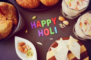 Assorted Indian Mahashivratri or Holi festival food like samosa, kachori, Kaju Katli, Puran Poli, dry fruits & Almond milk Thandai drink with Happy Holi wishes over black background with copy space.