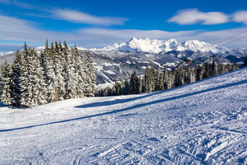 A winter landscape of a ski resort in Alps with hills, conifers, and mountains points covered in snow. A ski resort with a funicular, and ski tracks on a sunny morning with bright blue sky.