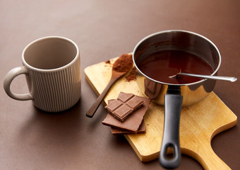 sweets, confectionery and culinary concept - pot with melted hot chocolate, cocoa powder in spoon, ceramic mug and wooden board on brown background