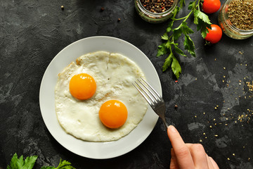 Woman eats fried eggs for breakfast, holds a fork. Fried eggs in a plate. Top view, dark background. Breakfast and ingredients.
