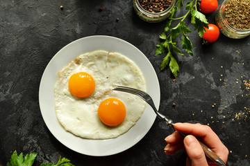 Woman eats fried eggs for breakfast, holds a fork. Fried eggs in a plate. Top view, dark background. Breakfast and ingredients.