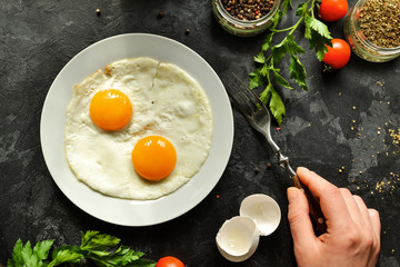 Woman eats fried eggs for breakfast, holds a fork. Fried eggs in a plate. Top view, dark background. Breakfast and ingredients.