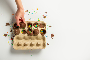 Female hand takes or holds chocolate eggs and multi-colored sweet candy decorations from a tray on a light background. Easter concept, easter treats, bunny. Banner. Flat lay, top view
