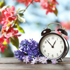 Black alarm clock and flowers on wooden table against blurred background. Spring time