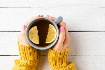 Overhead shot of female hands knitted sweater holding cup of tea with lemon on white wooden table