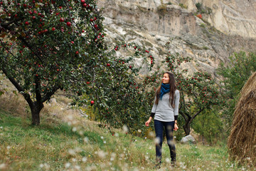 Young beautiful woman walking elegant among apple trees with big red apples in garden. Background of rocky mountains