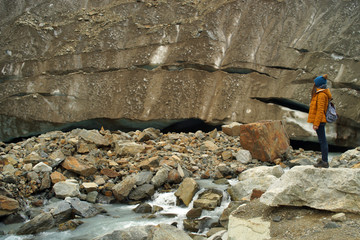 Young woman hiker in warm clothes with backpack standing on stones in front of glacier and mountain, view from side