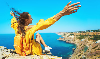 Close up portrait of taned traveler woman sitting on top rock beach with raised hands, bright...