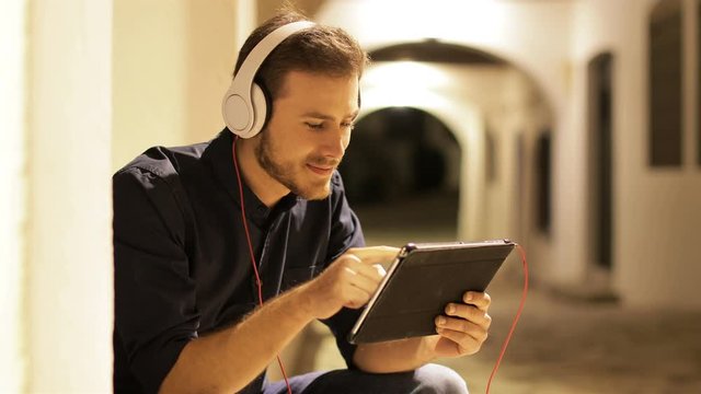 Happy man browsing on tablet wearing headphones sitting in the street in the night