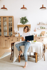 Young stylish girl in a white T-shirt sits on a table in her cozy kitchen with a laptop in her hands